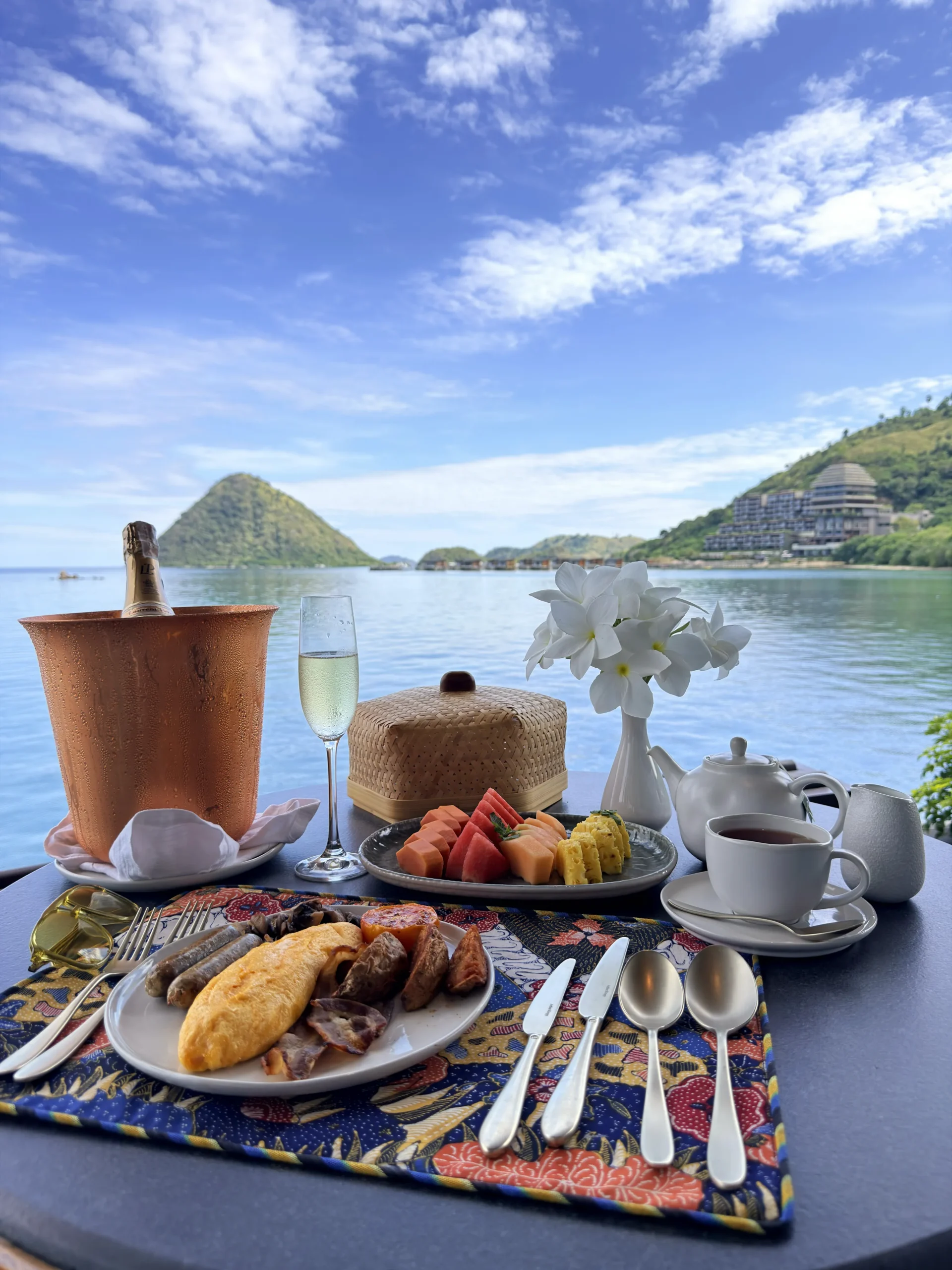 A gourmet in-villa breakfast spread set on a private outdoor terrace, framed by the iconic panoramic views of the Flores Sea