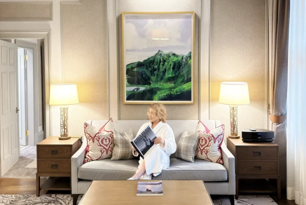 A person, attributed as Stefano Malachi, in a white bathrobe and slippers, is seated on a plush velvet sofa within a grand hotel suite, reading a book titled 'The Planets'. The scene unfolds in the opulent living area of the Somerville Suite at InterContinental Edinburgh The George, featuring a large framed Scottish highland painting above the sofa, paneled walls, elegant lamps, textured armchairs, a detailed coffee table with art books, and decorative pillows with a Scottish design.