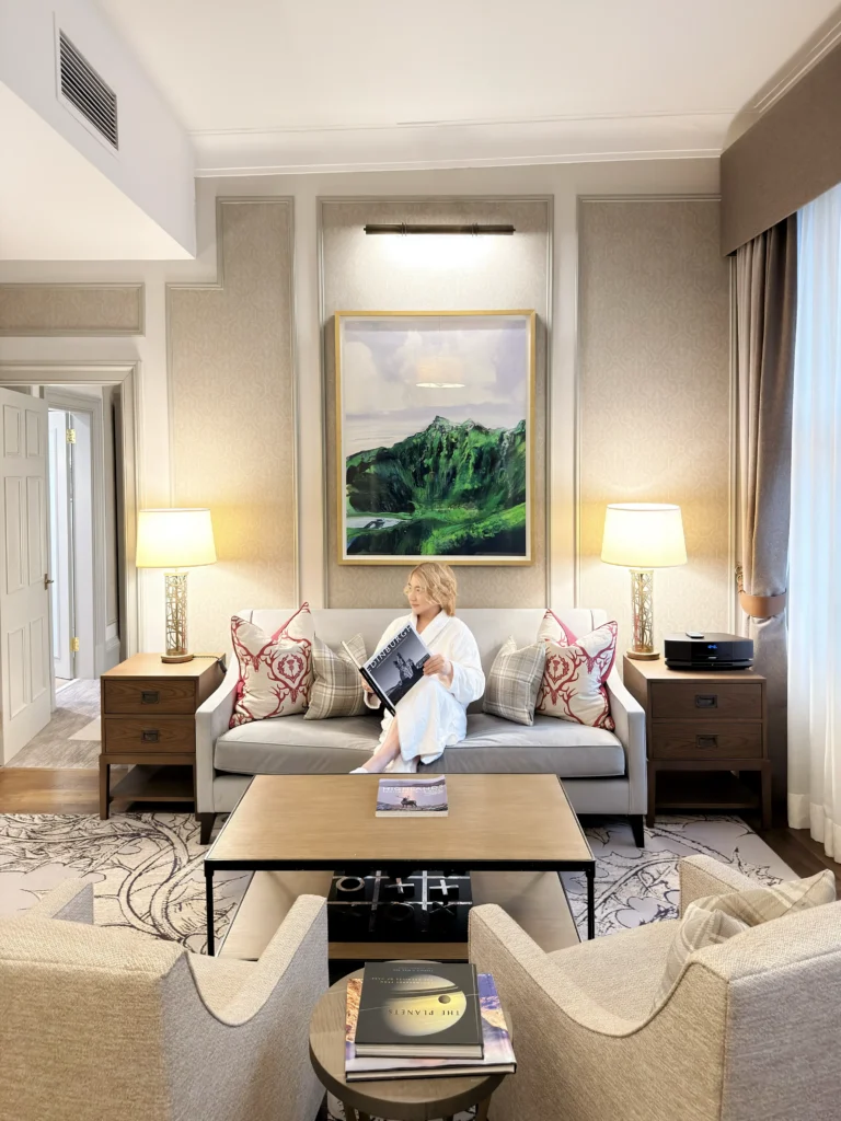 A person, attributed as Stefano Malachi, in a white bathrobe and slippers, is seated on a plush velvet sofa within a grand hotel suite, reading a book titled 'The Planets'. The scene unfolds in the opulent living area of the Somerville Suite at InterContinental Edinburgh The George, featuring a large framed Scottish highland painting above the sofa, paneled walls, elegant lamps, textured armchairs, a detailed coffee table with art books, and decorative pillows with a Scottish design.