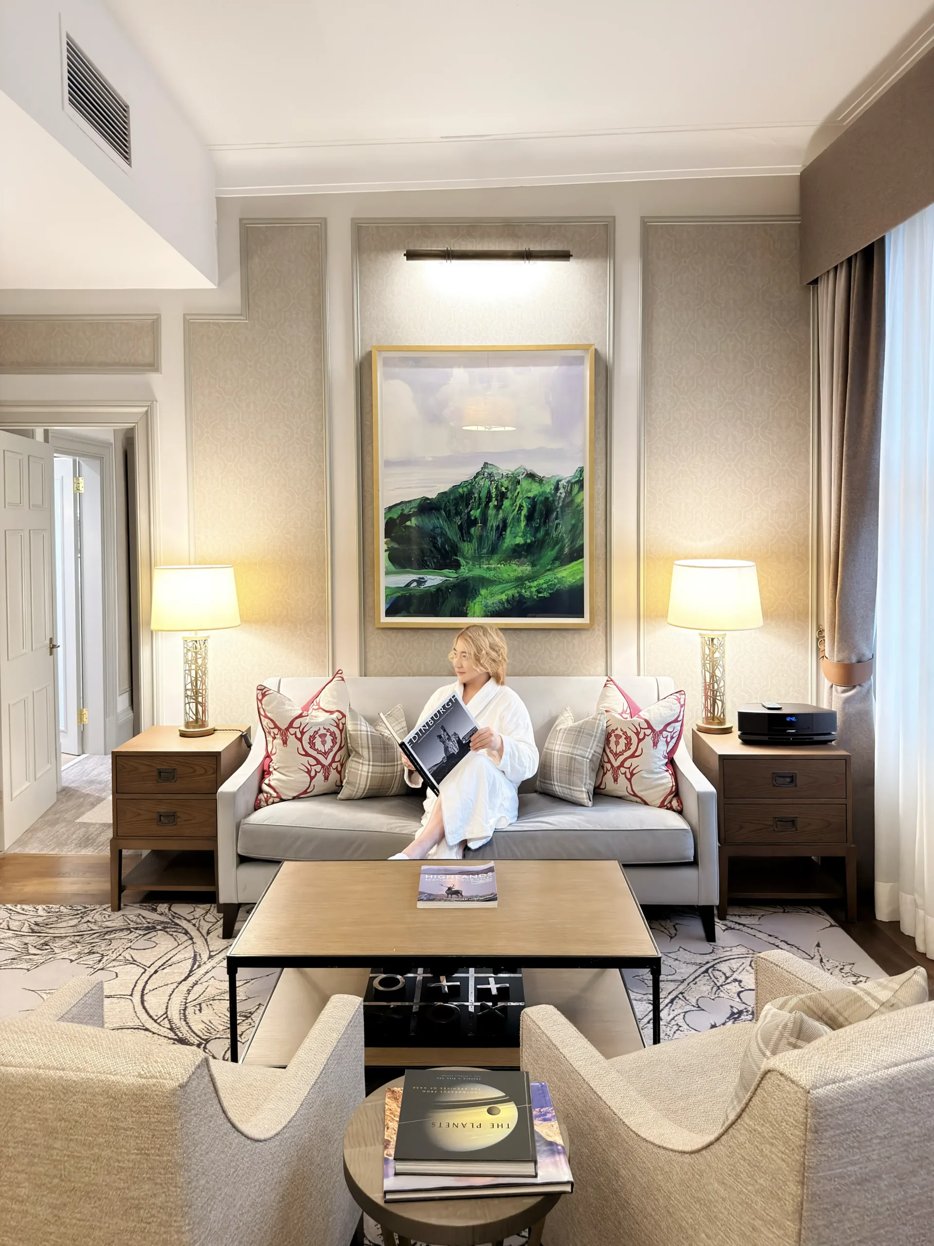 A person, attributed as Stefano Malachi, in a white bathrobe and slippers, is seated on a plush velvet sofa within a grand hotel suite, reading a book titled 'The Planets'. The scene unfolds in the opulent living area of the Somerville Suite at InterContinental Edinburgh The George, featuring a large framed Scottish highland painting above the sofa, paneled walls, elegant lamps, textured armchairs, a detailed coffee table with art books, and decorative pillows with a Scottish design.