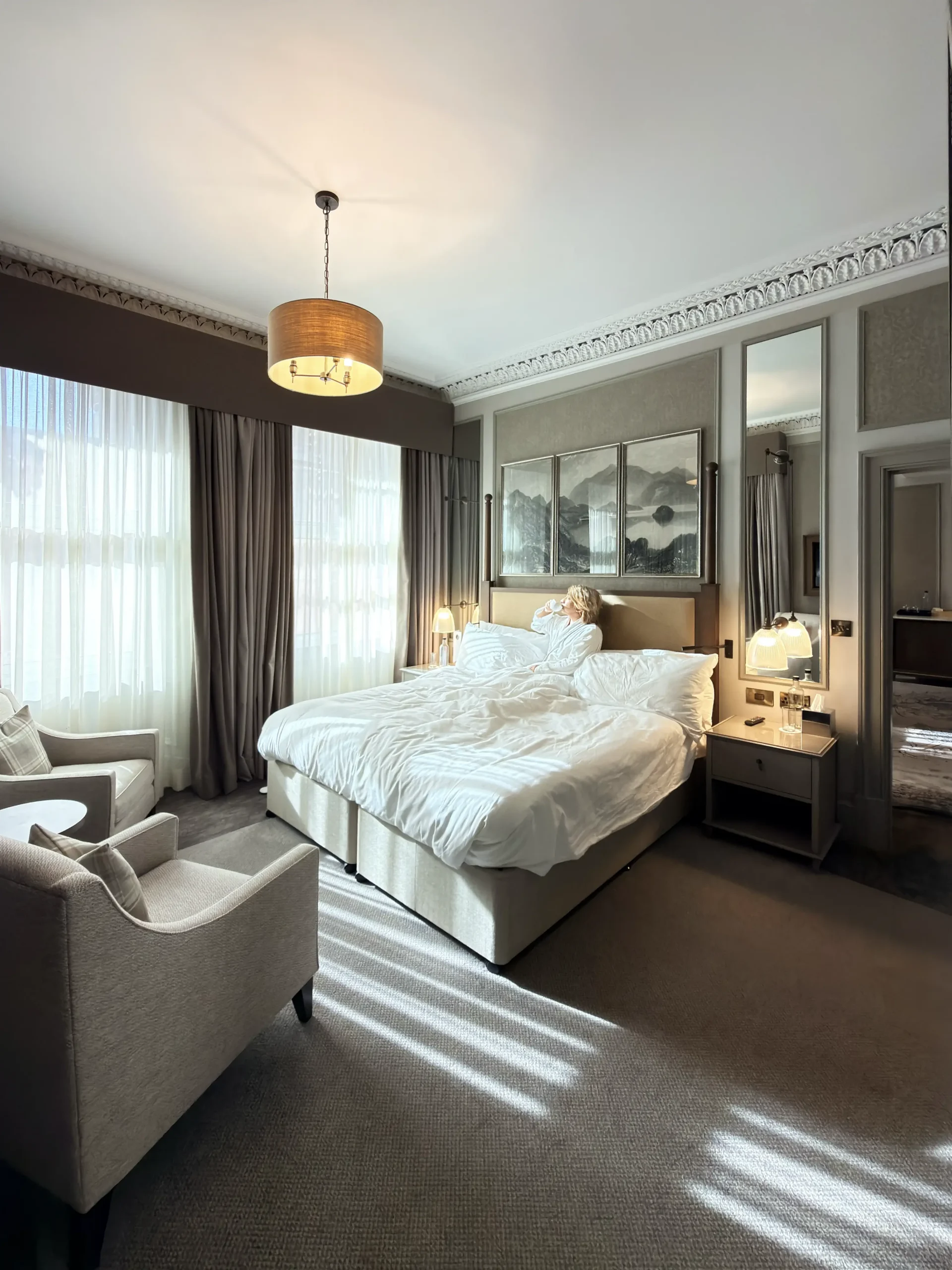 Stefano Malachi, in a plush white bathrobe, sits comfortably in a large, sun-dappled king-size bed in the luxurious Somerville Suite, InterContinental Edinburgh The George. She is holding a mug and gazing towards the light, enjoying a quiet morning moment. Sunlight streams through tall windows, casting light streaks on the grey carpet and illuminating the elegant, high-ceilinged room with its traditional crown molding, paneled walls, and a large landscape triptych.