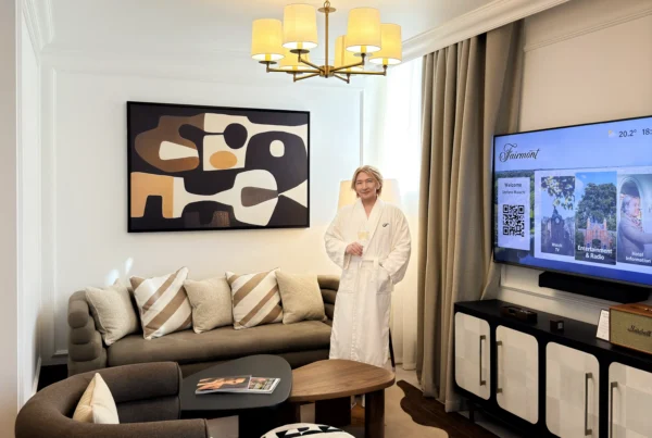 Stefano Malachi stands comfortably in a luxurious Fairmont Cheshire The Mere hotel suite, wearing a white bathrobe and holding a glass of champagne. The room features a mid-century modern aesthetic with a large abstract painting, unique seating, and a TV displaying the "Fairmont" welcome screen