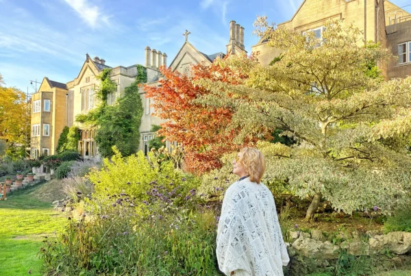 Stefano Malachi stands in full profile wearing a long, flowing white lace knit cardigan over patterned trousers, looking toward the lush gardens of The Bath Priory during golden hour, with the historic honey-colored stone country house and colorful autumn trees bathed in afternoon light beneath a vibrant blue sky