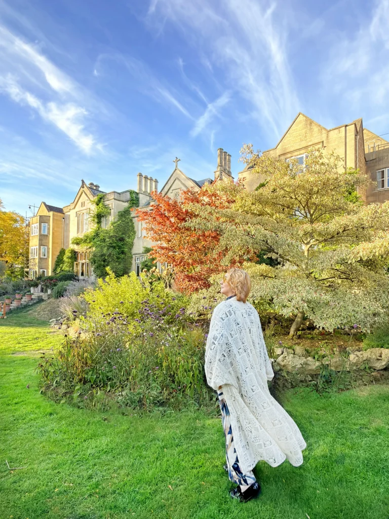 Stefano Malachi stands in full profile wearing a long, flowing white lace knit cardigan over patterned trousers, looking toward the lush gardens of The Bath Priory during golden hour, with the historic honey-colored stone country house and colorful autumn trees bathed in afternoon light beneath a vibrant blue sky