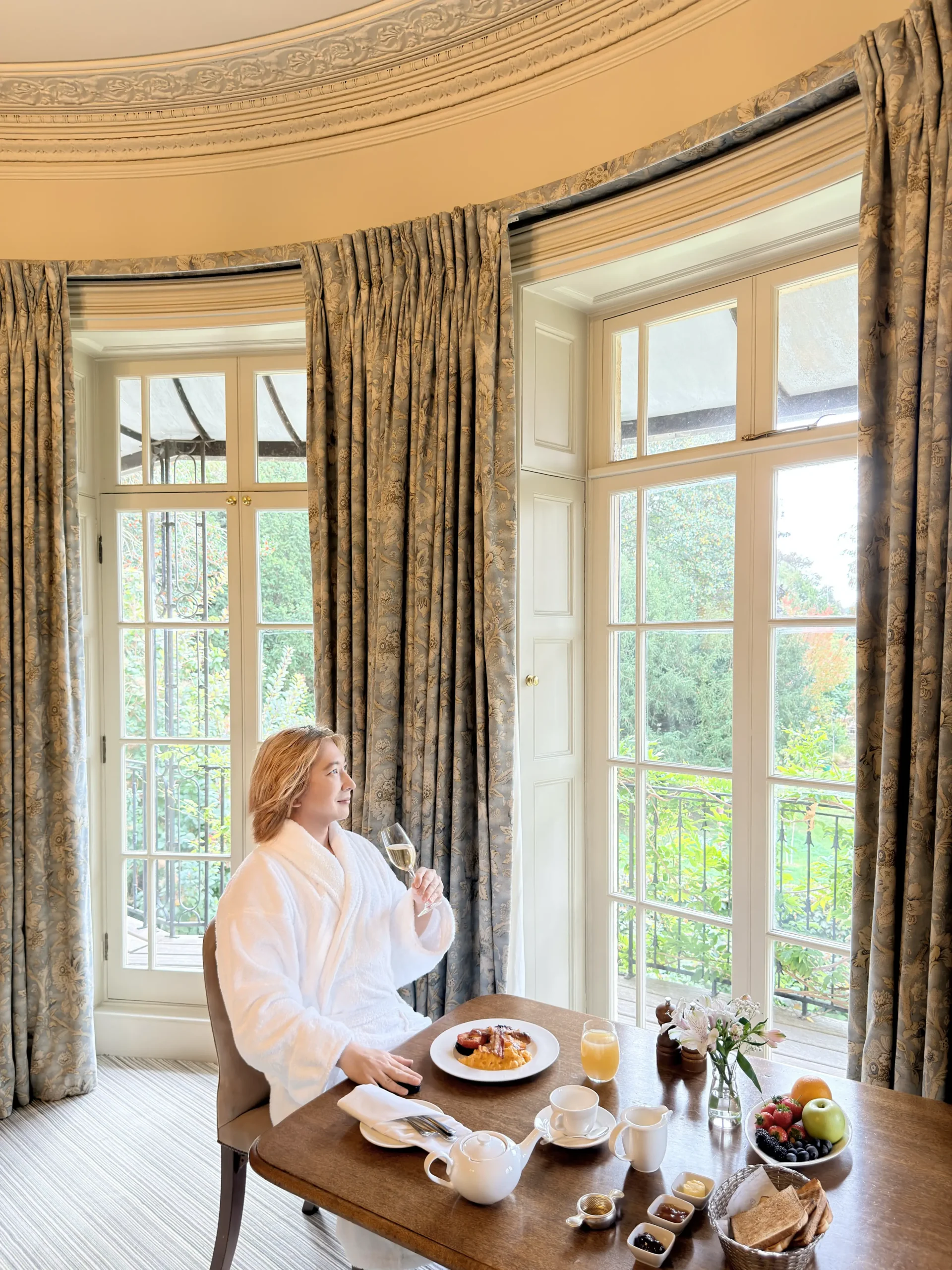 Stefano Malachi, wearing a plush white hotel bathrobe, sits at a wooden dining table enjoying a luxury in-room breakfast with champagne, scrambled eggs, pastries, fresh fruit, and tea in a bay window alcove at The Bath Priory, looking out toward garden views.