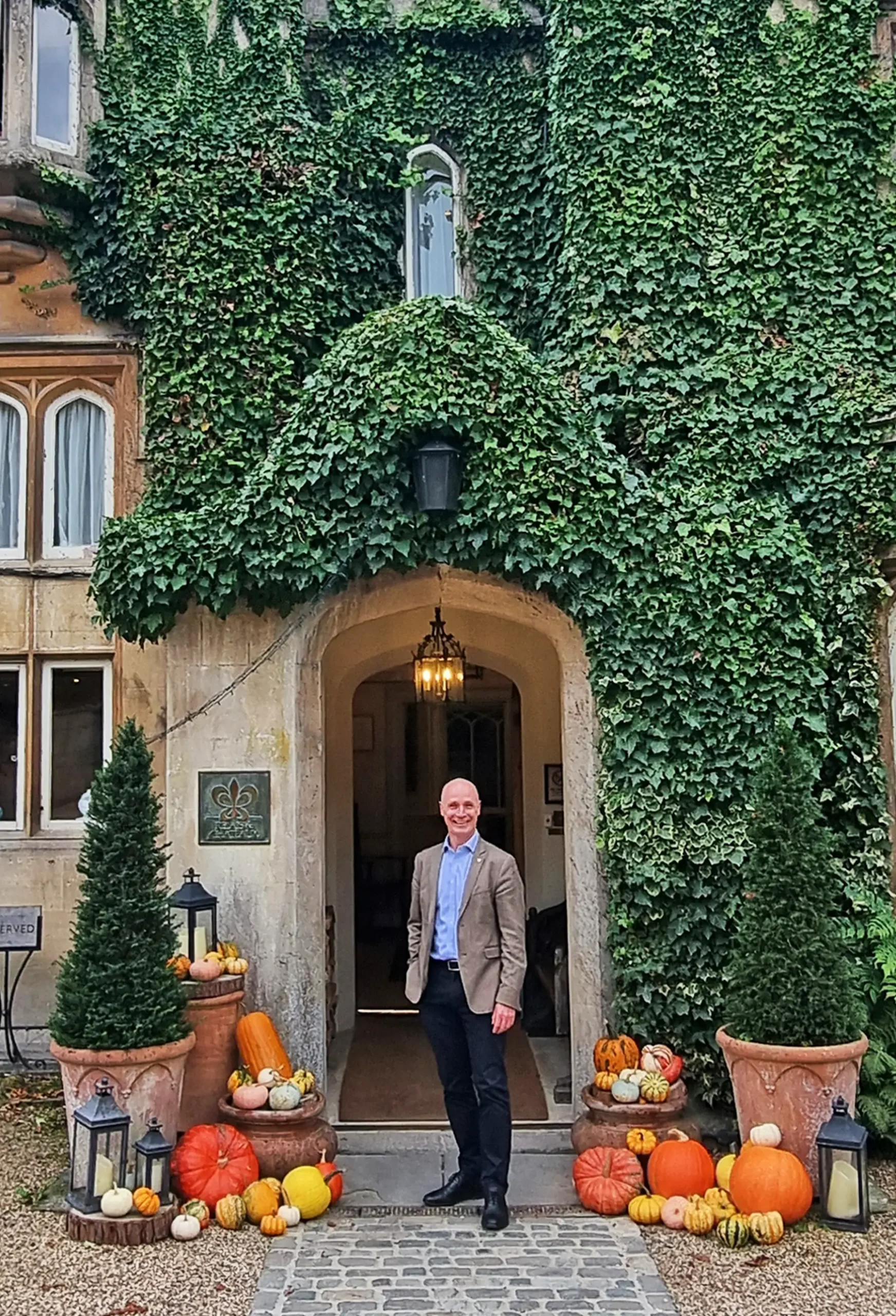 Gurval Durand, Regional General Manager of The Bath Priory, stands smiling in a sharp tailored blazer at the arched entrance of the historic ivy-covered stone hotel, flanked by festive autumnal displays of pumpkins, gourds, potted topiary trees, and lanterns.