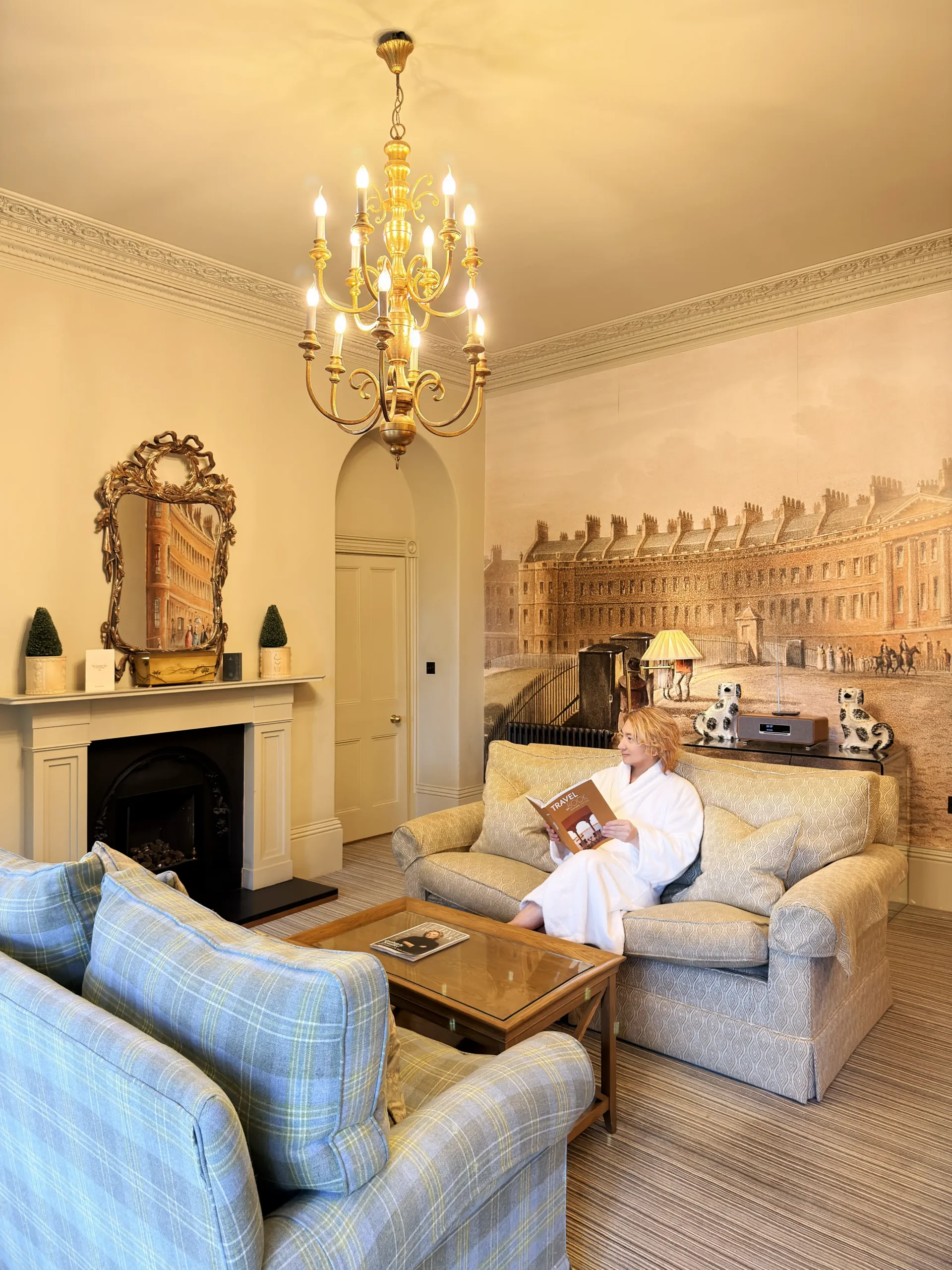 Stefano Malachi, dressed in a plush white bathrobe, sits comfortably on a patterned sofa reading a book in the Lansdown Suite at The Bath Priory. The room features a grand brass chandelier, an ornate gilded mirror above a classic fireplace, plush plaid armchairs, and a striking floor-to-ceiling mural depicting historic Georgian architecture.