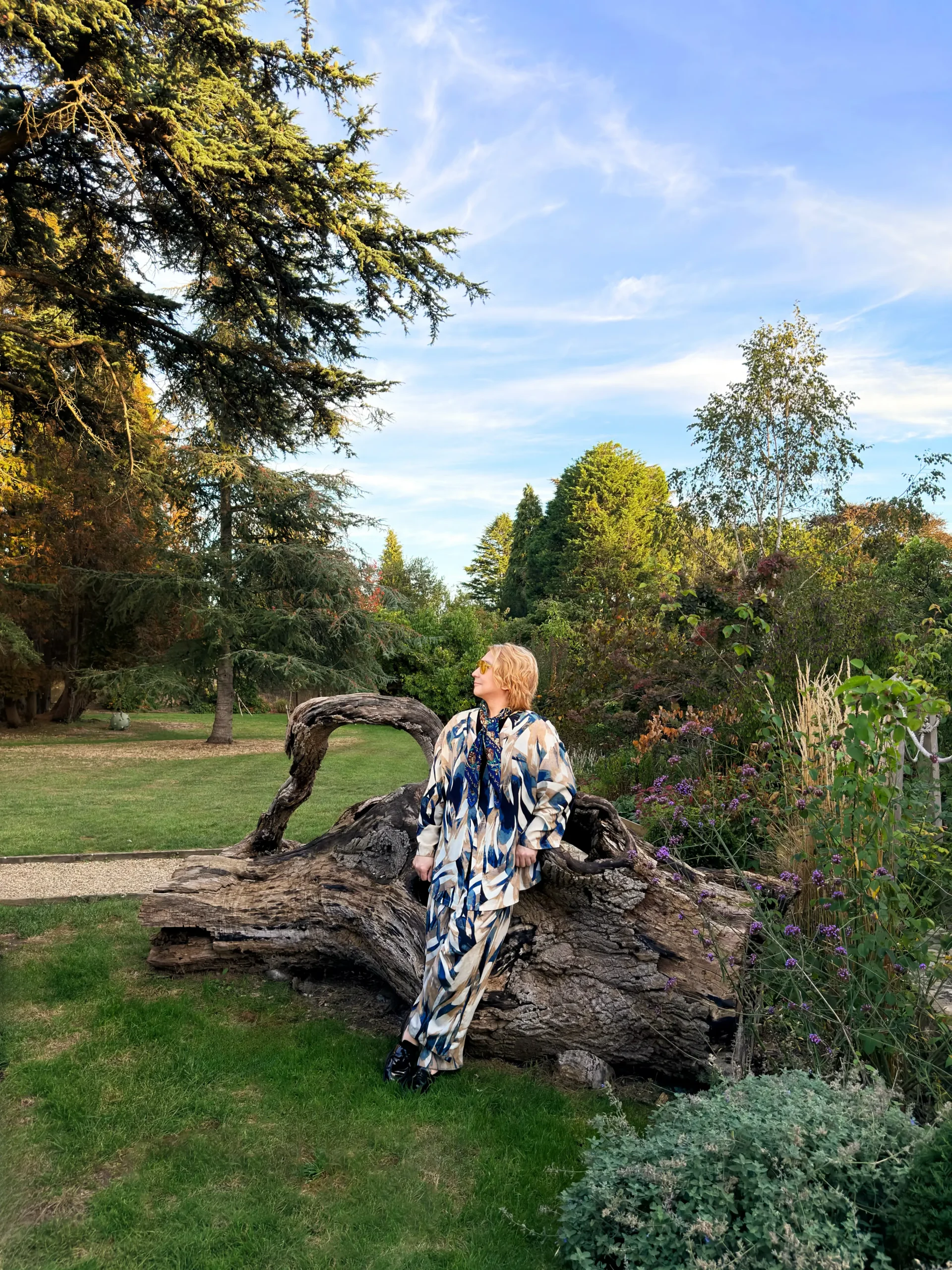 Stefano Malachi, wearing a stylish blue-and-beige abstract patterned silk co-ord set with sunglasses and a silk neck scarf, leaning casually against a large, sculptural fallen tree trunk in the lush gardens of The Bath Priory under a clear blue sky.